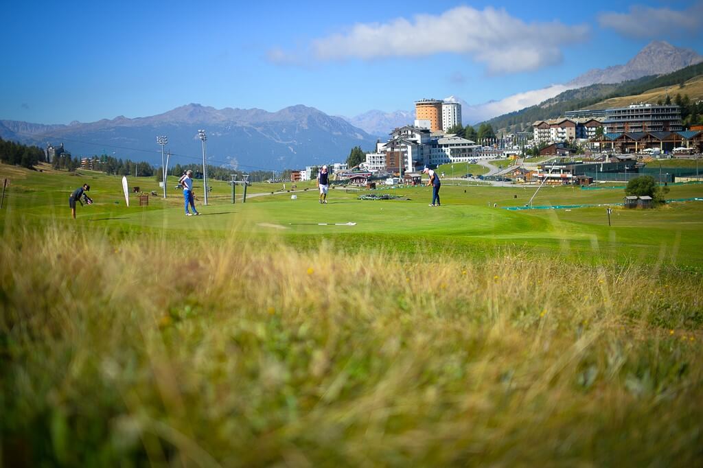 Four golfers play on a manicured green with mountains and buildings in the background, surrounded by flowers and grass.